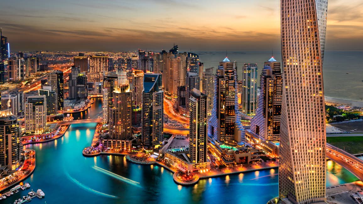 A mesmerizing aerial view of the Dubai skyline and coastline bathed in the warm hues of a sunset, with the city's dazzling lights on display during the Dubai International Boat Show.